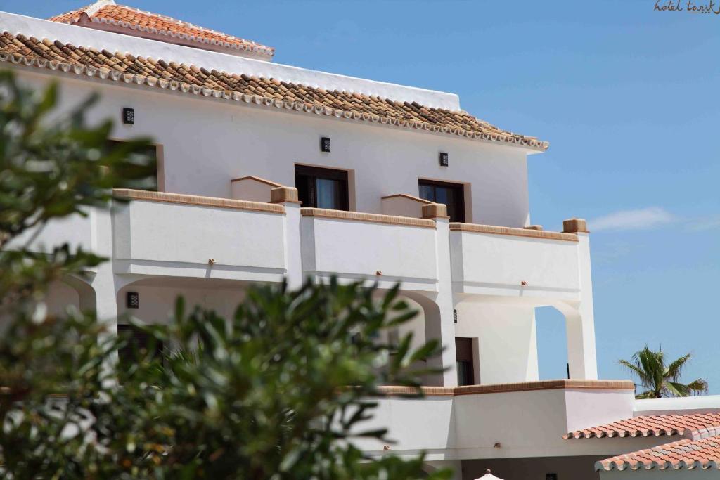 Viesnīcas ēka ar baltiem sienām un bruģētu jumtu, apzaļumota ar zālēm un palmām. Skats uz terasi un saulainu debesi.