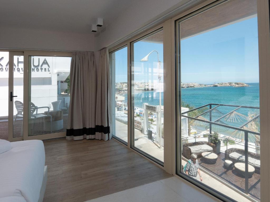 Skats no viesnīcas numura uz jūru un pludmali, ar gaišām krāsām un mūsdienīgu interjeru. Logi ļauj ienākt dabiskajai gaismai, radot relaksējošu atmosfēru.