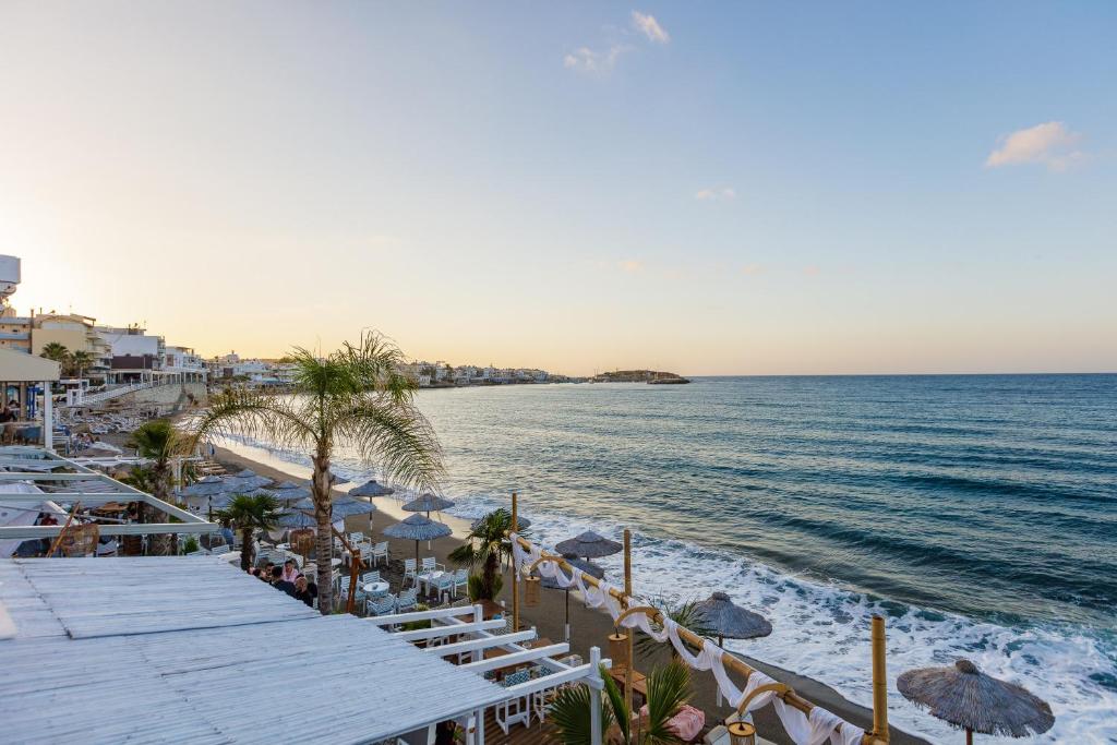 Pludmales ainava ar saulrietu, kurā redzami palmas, sauļošanās krēsli un saulessargi, kas piekraste ir klāta ar smiltīm. Kluss jūras skats ar viļņiem un ēkām fona.