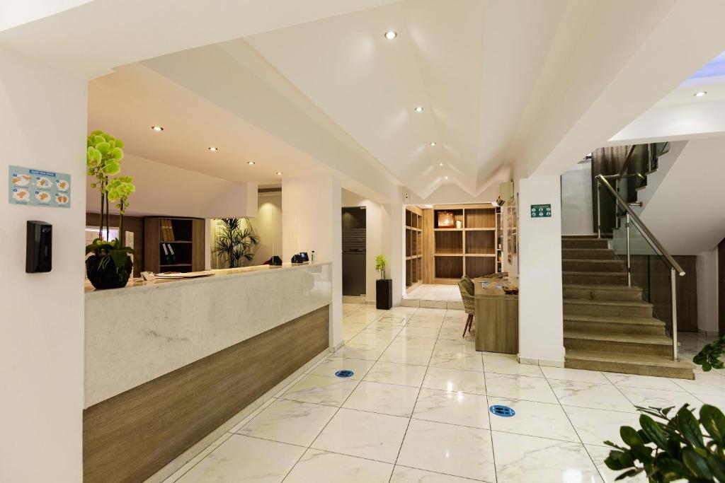 Mūsdienīga viesnīcas recepcija ar gaišām flīzēm, koka akcentiem un augiem, radot patīkamu un viesmīlīgu atmosfēru. Redzama kāpņu telpa un uzņemšanas zona.