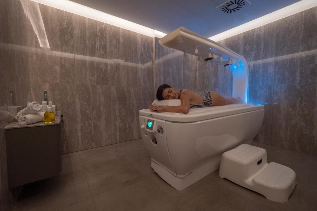 Relaksējoša vannas procedūra modernā spa vidē ar mūsdienīgu vannu un apgaismojumu. Blakus redzami dvieļi un kosmētika.