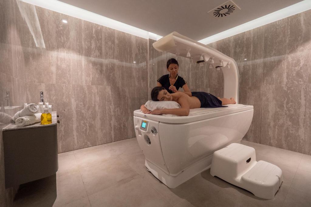 Mūsdienīga spa telpa, kur masāžas terapeits sniedz relaksējošu masāžu klientam, kas guļ uz modernā masāžas galda. Telpai ir eleganta interjers ar akmens apdari un maigu apgaismojumu.