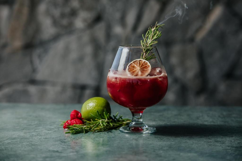 Sarkanais kokteilis ar citronu un rozmarīnu, papildināts ar laimu un avenēm, uz tumša fona.