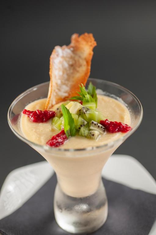 Krēmveida deserts pasniegts martini glāzē, rotāts ar augļu gabaliņiem un kraukšķīgu cepumu.