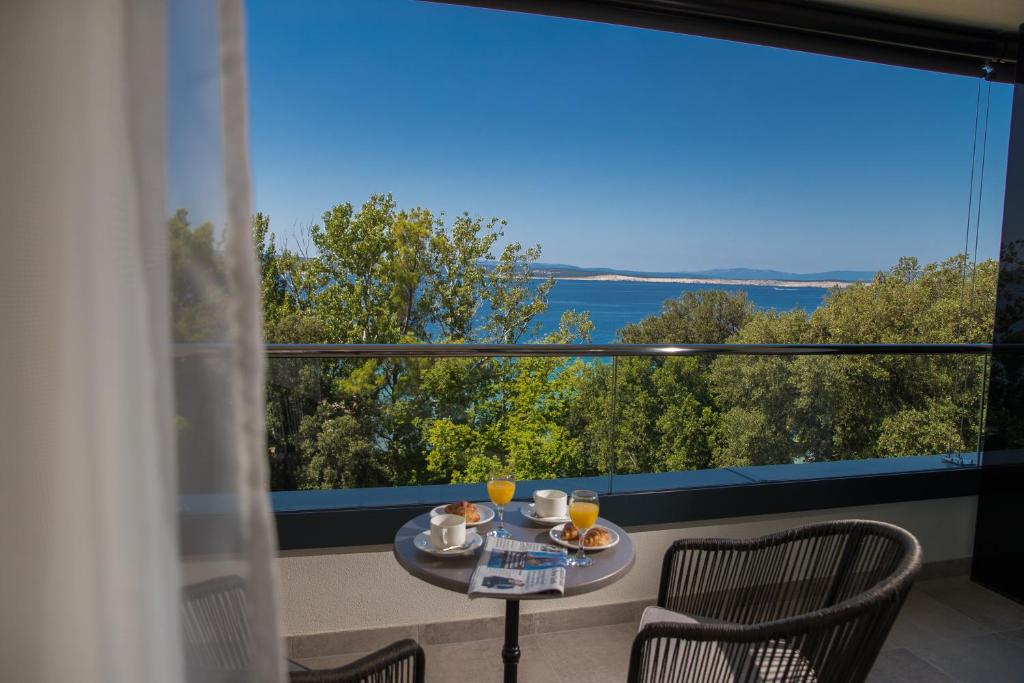 Balkona skats uz jūru ar brokastīm uz galda, kur ir kafija, sula un uzkodas. Zaļumi un zila jūra veido mierīgu atmosfēru.