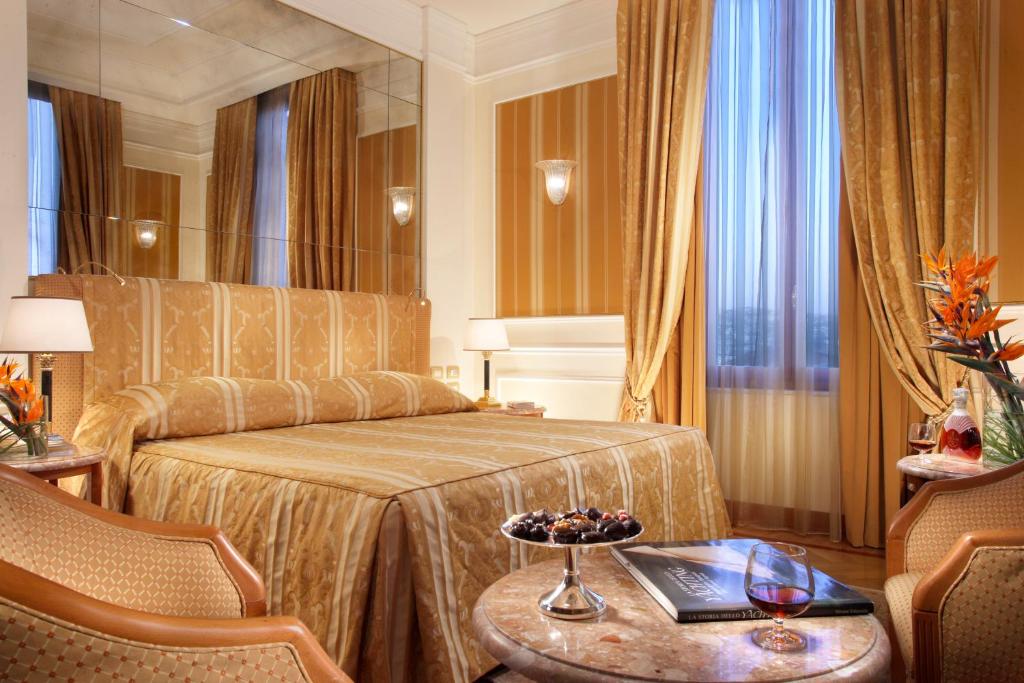 Elegants viesnīcas numurs ar dzeltenām un zeltainām tonām, ērta gulta un mūsdienīgs interjers. Blakus atrodas kumode ar našķiem un grāmatu.