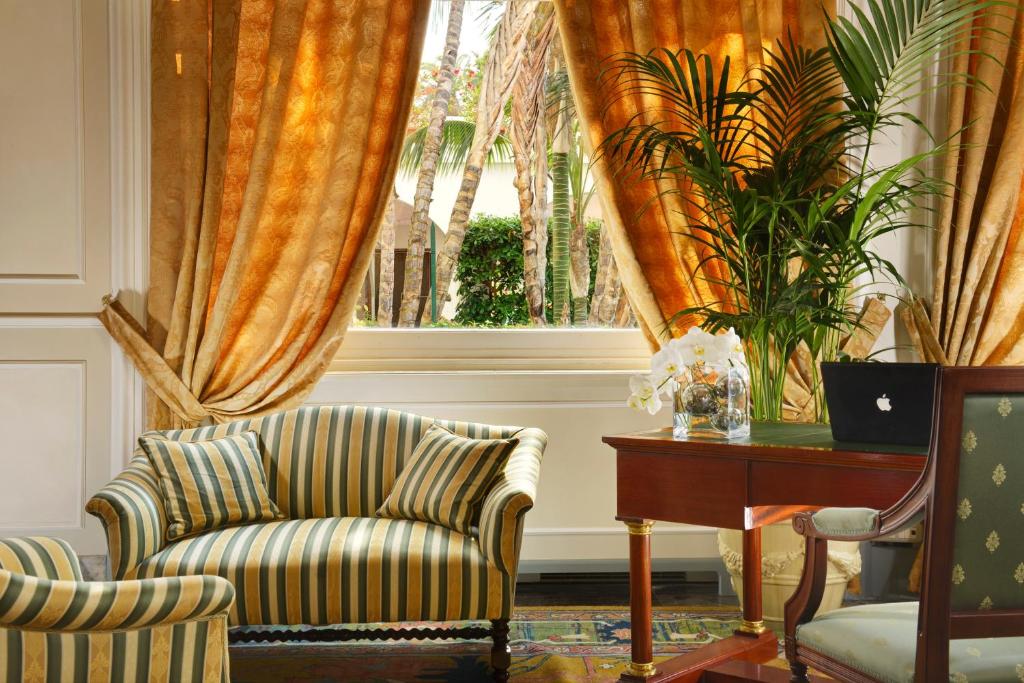 Elegantā viesistaba ar dzeltenām aizkarām, svītrainu dīvānu un galdu, uz kura novietots klēpjdators un ziedi. Logs paver skatu uz zaļajiem augiem ārpusē.