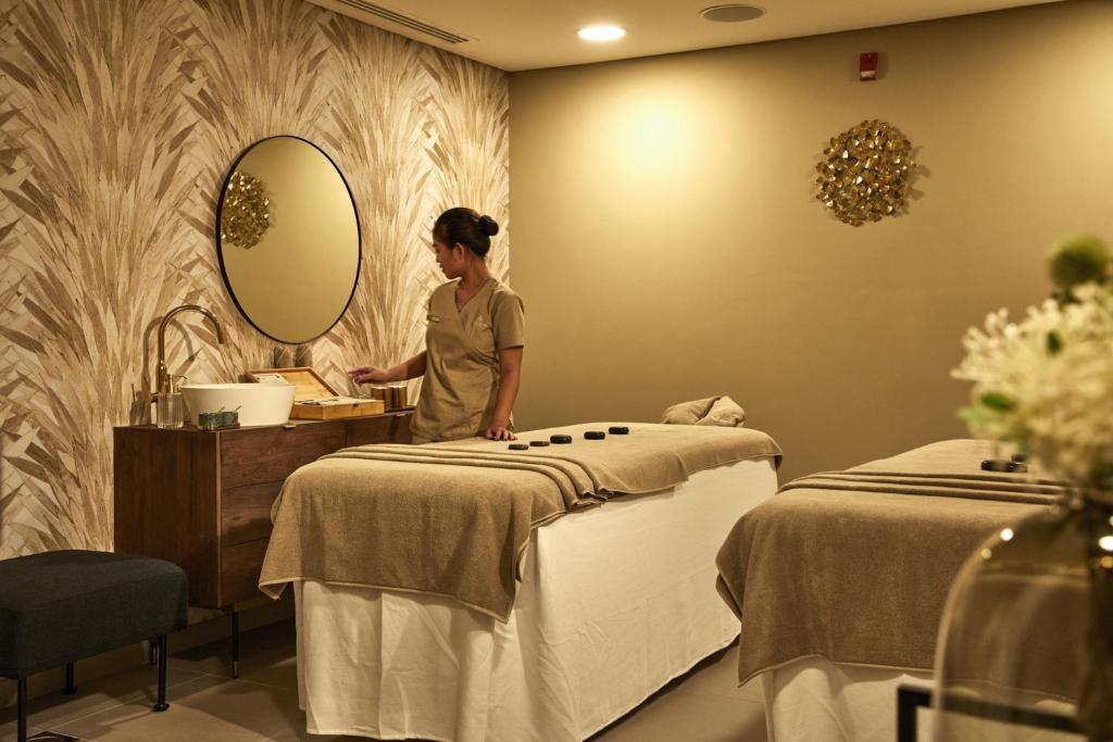 Mūsdienīgs SPA interjers ar masāžas galdiem, apgaismojumu un relaksējošu atmosfēru. Darbiniece sagatavo masāžas rīkus, lai nodrošinātu klientu komfortu.