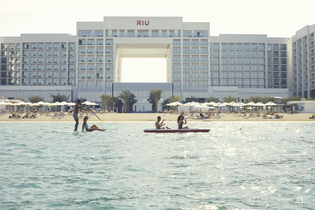 Pludmales ainava ar viesnīcu RIU fona, kur cilvēki nodarbojas ar ūdens sporta aktivitātēm, piemēram, kanoe un stand-up paddleboarding.