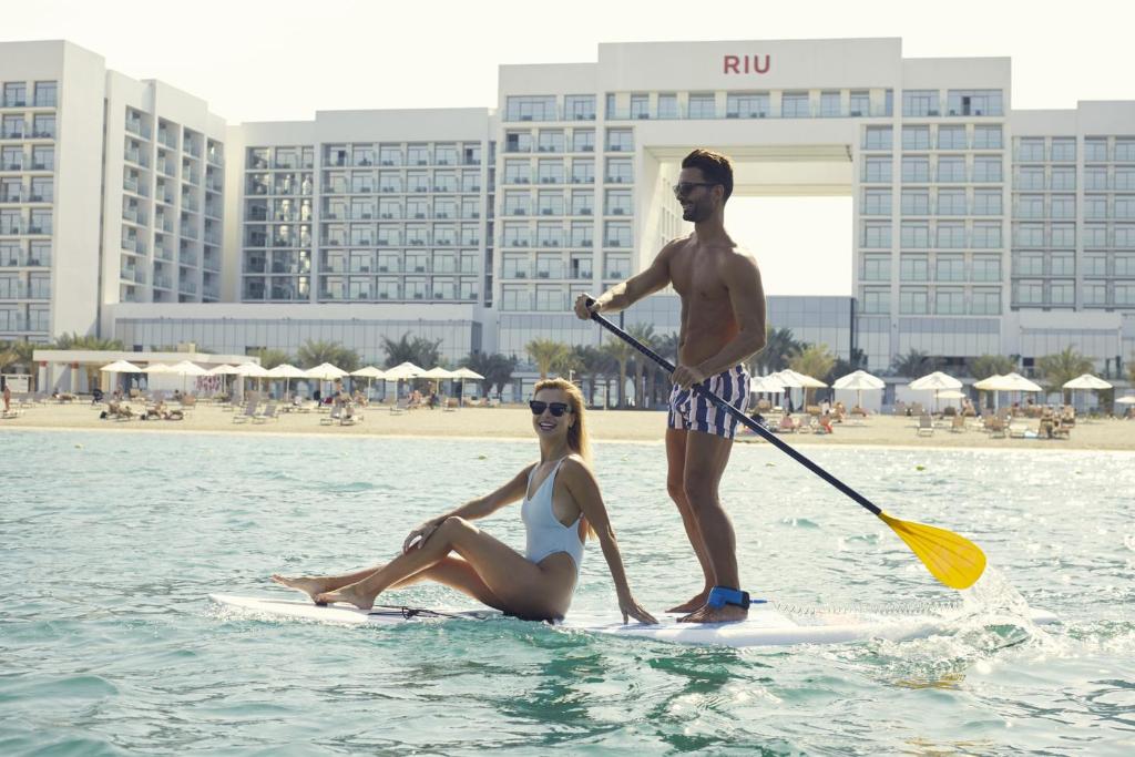 Pārgājiens ar stand up paddle dēli, kurā redzams pāris ar saulesbrillēm, fons ir pludmale un viesnīca Riu. Ideāli piemērots vasaras atpūtai.