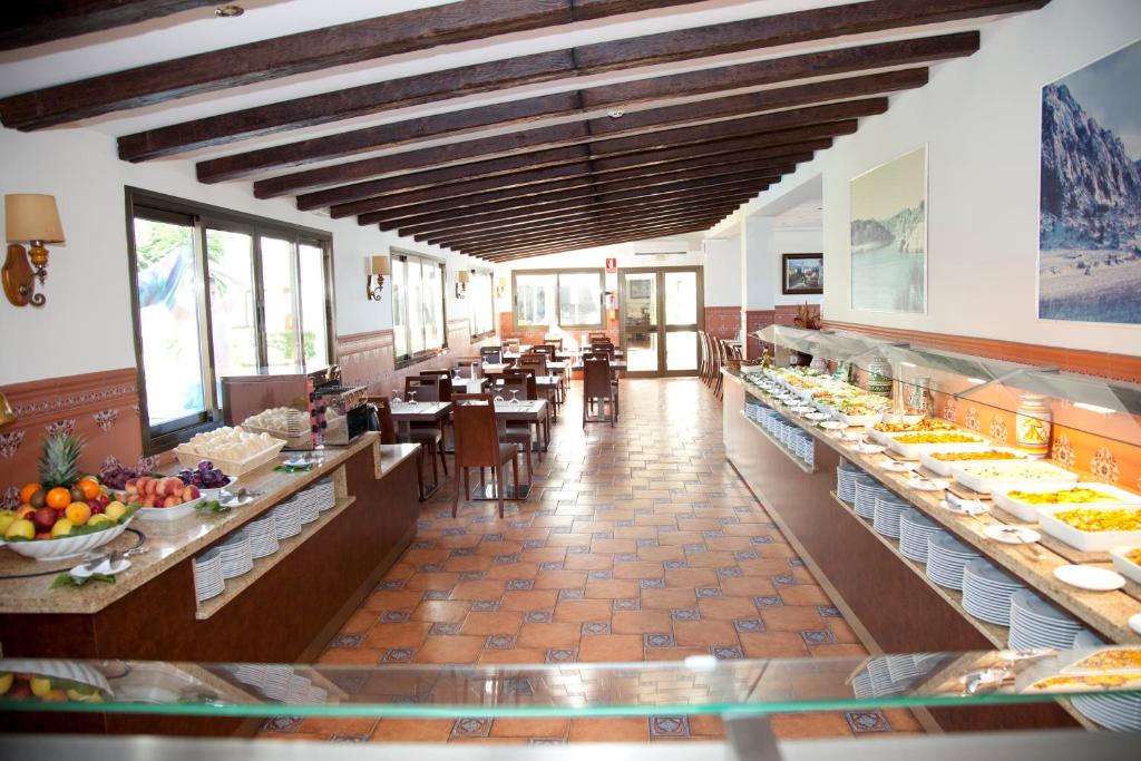 Restorāna iekšējais skats ar ēdamgaldiem un bagātīgu bufeti, kas piedāvā augļus un dažādus ēdienus. Gaisotne ir gaiša un mājīga, ar koka sijām un flīžu grīdu.