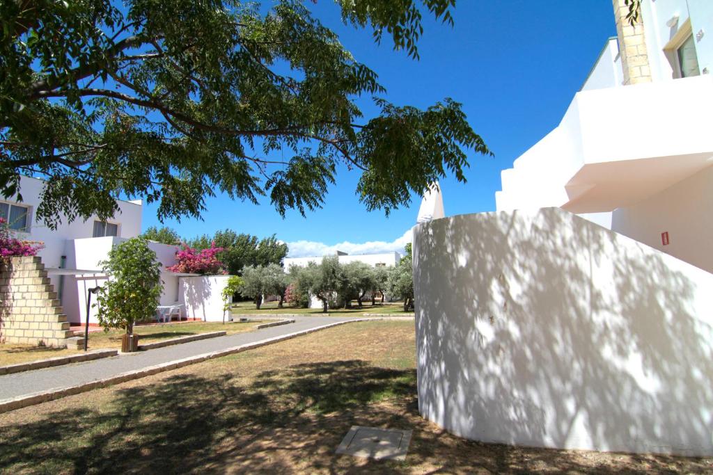 Mūsdienīgs atpūtas komplekss ar baltiem ēku fasādēm un zaļām ainavām, kurā redzami koki un ziedi, kas veido patīkamu atmosfēru. Ideāla vieta relaksācijai un atpūtai.