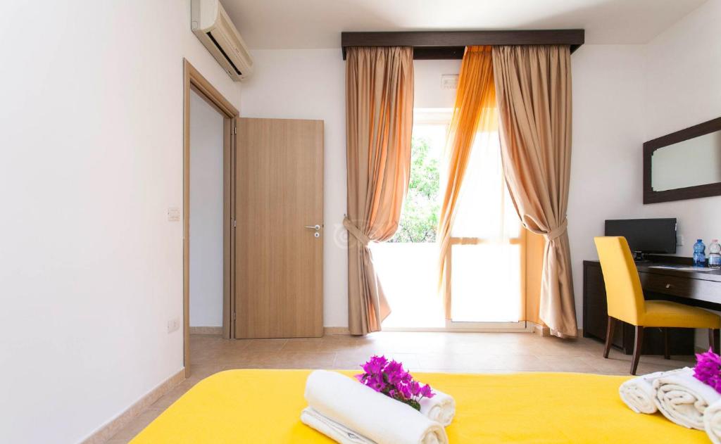 Gaišs un moderns guļamistabas interjers ar dzeltenu gultasveļu, izrotātu ar ziediem, un ērtiem mēbelēm, kas ietver rakstāmgaldu un krēslu. Logi ar viegliem aizkariem nodrošina dabīgu apgaismojumu.