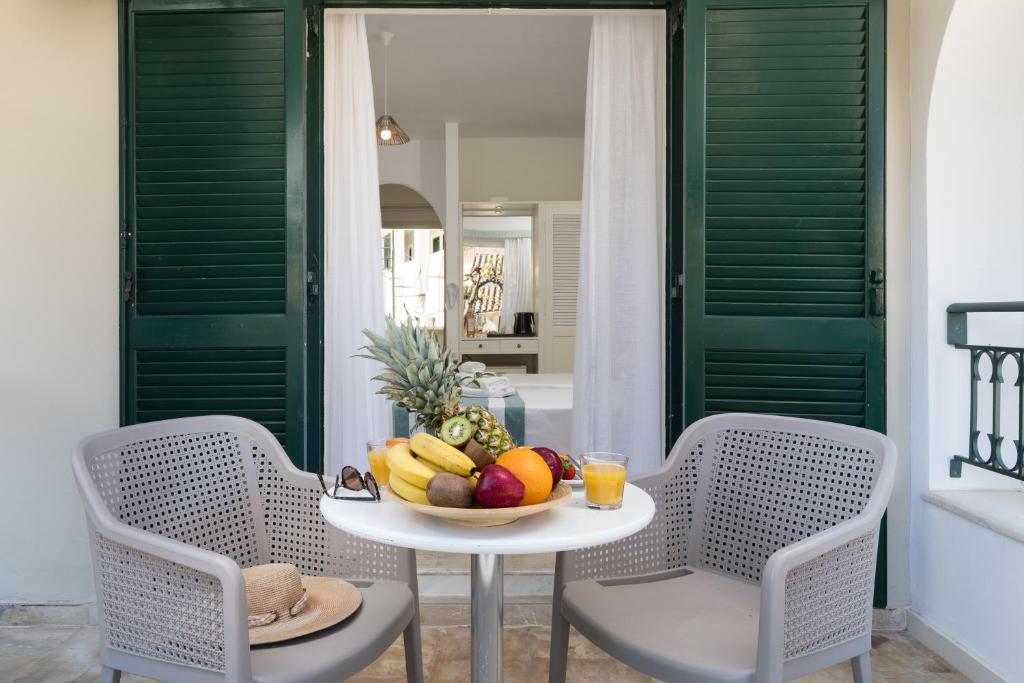 Balcony with two modern chairs and a round table, adorned with a vibrant fruit platter and a glass of juice, overlooking a cozy interior with green shutters and soft lighting.