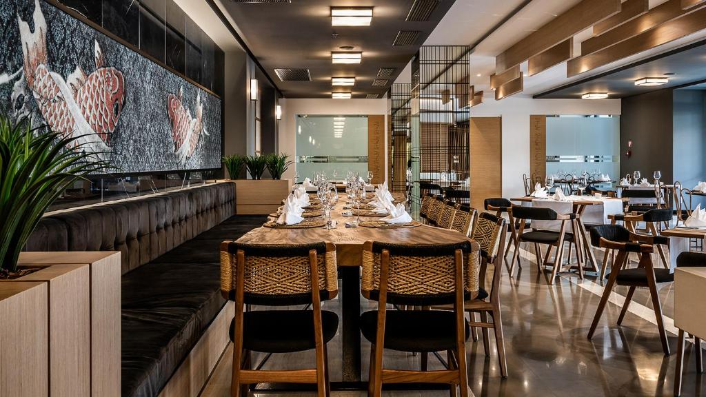 Mūsdienīgs restorāns ar elegantu interjeru, kurā ir liels koka galds un izsmalcinātas galda klāšanas detaļas. Uz sienas redzama mākslinieciska zivs mozaīka, kas piešķir telpai unikālu atmosfēru.