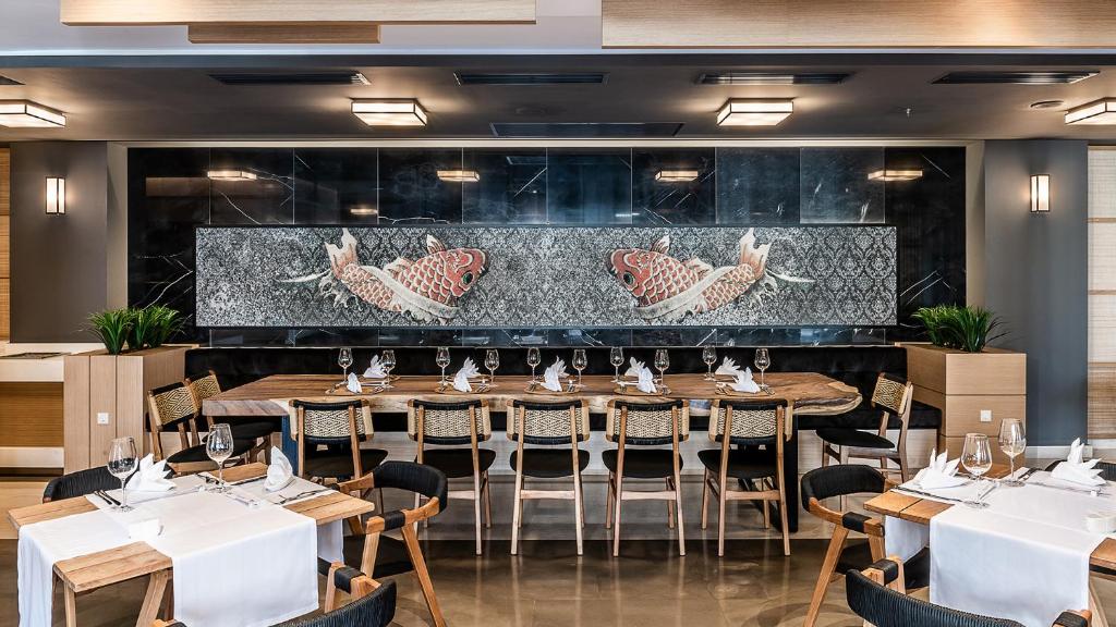 Mūsdienīga restorāna iekārtojums ar elegantu galdu, kura centrā ir krāšņa mozaīka ar zivīm. Atmosfēra ir silta un aicinoša, ideāli piemērota ēdienreizei kopā ar draugiem vai ģimeni.