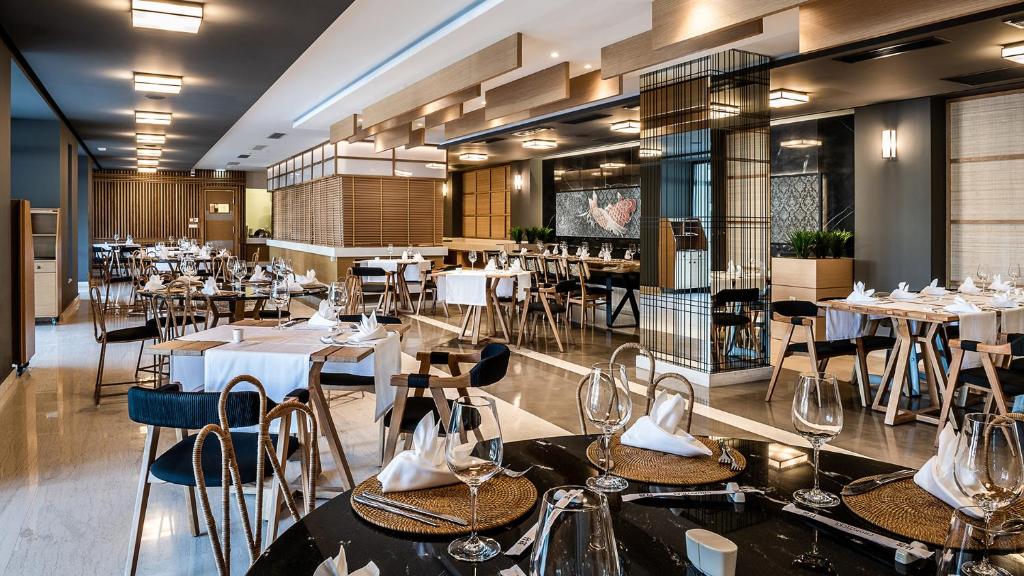 Mūsdienīgs restorāns ar elegantu interjeru, kas ietver galdiņus ar galda piederumiem un gleznainiem dekoratīviem elementiem. Telpa ir gaiša un plaša, radot patīkamu atmosfēru.