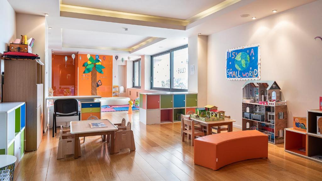 Bērnu rotaļu istaba ar krāsainiem mēbeļu elementiem, galdiem un rotaļlietām, kas veicina radošumu un spēlēšanos. Sienas rotā bērnu zīmējumi un uzraksti, radot draudzīgu un jautru atmosfēru.