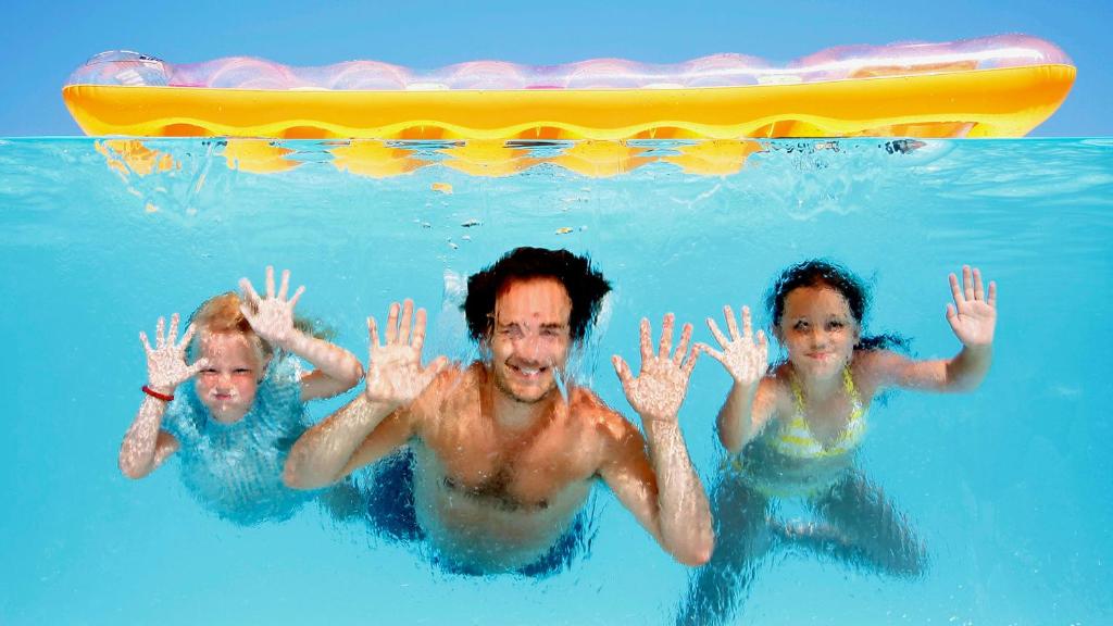 Trīs cilvēki, tostarp divi bērni un pieaugušais, priecīgi peld ūdenī, rādot rokas uz augšu. Virs tiem redzama dzeltena peldoša matrace.