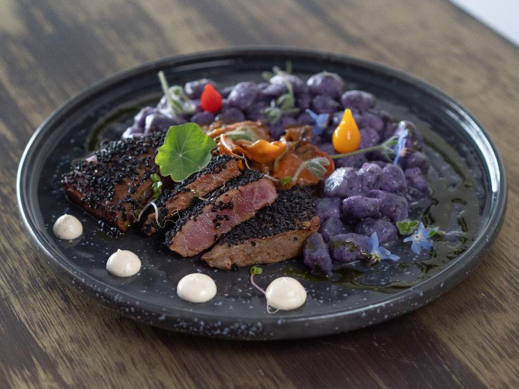 Melnā sezama garšviela ceptai gaļai ar purpura gnocchi un krāsainiem dārzeņiem, pasniegts elegantā melnā šķīvī.