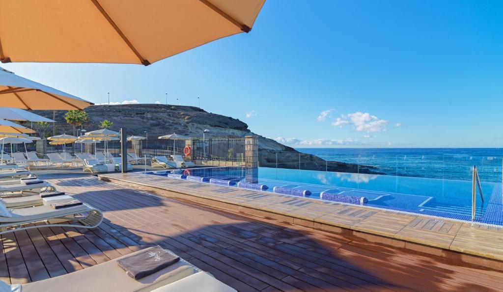 Luksusīgs baseins ar skatu uz jūru, apņemts ar sauļošanās krēsliem un saulessargiem, kas atrodas pie pludmales. Skats uz klinšainu krastu un zilo debesu fona.