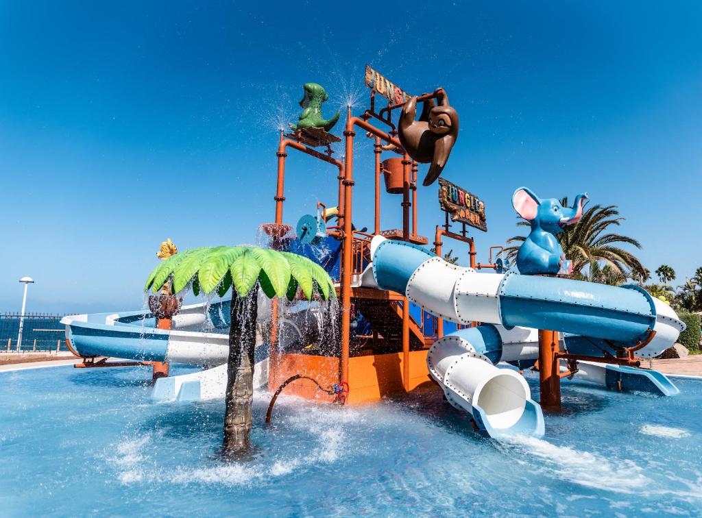 Ūdens atrakciju parks ar krāsainām slidkalniņiem un dzīvnieku figūrām, piemēram, ziloņu un lauvām, zem skaidras debess.