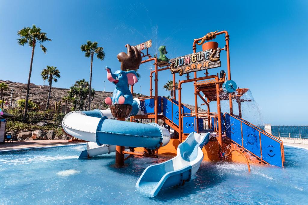 Ūdens slidkalniņš tropu tematiskajā parkā ar zilo ziloņu figūru un ūdens strūklām, iepriecinot apmeklētājus saulainā dienā.