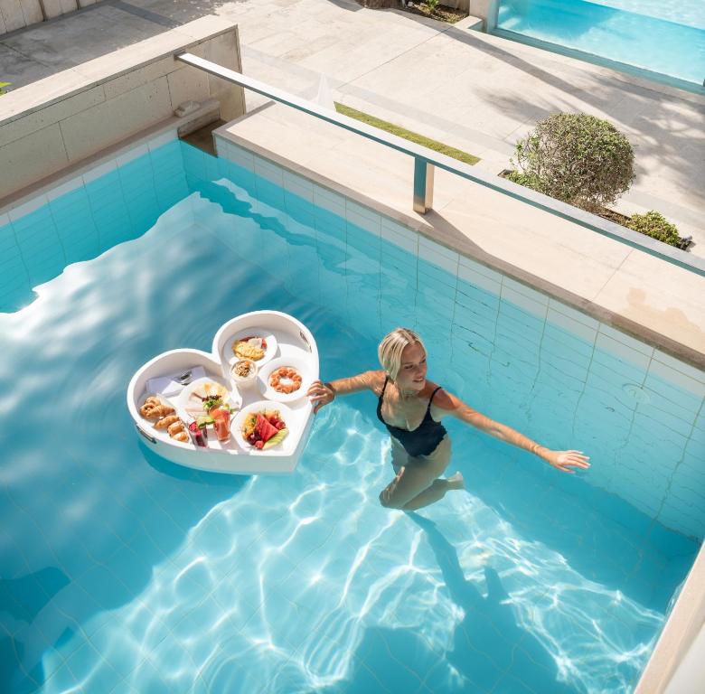 Sieviete peldbaseinā baudīja ēdienus no sirds formas šķīvja. Ūdens ir dzidrs un saule spīd, radot relaksējošu atmosfēru.