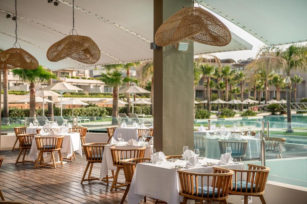 Elegants restorāns ar koka mēbelēm un baltiem galdautiem, kas piedāvā skatu uz peldbaseinu un palmām. Gaismas ķermeņi no dabīgiem materiāliem piešķir siltu atmosfēru.