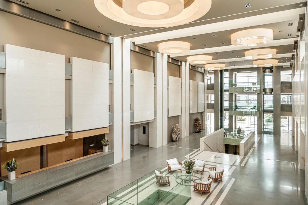 Mūsdienīgs un plašs viesnīcas vestibils ar augstiem griestiem, elegantiem lampām un modernu interjeru. Telpā redzami mēbeļu elementi un dekoratīvi augi, kas piešķir atmosfēru.