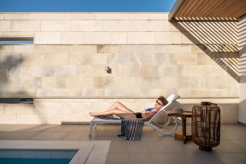 Atpūtas brīdis pie baseina, kurā cilvēks guļ uz sauļošanās krēsla, baudot sauli. Apkārt ir moderns interjers ar dabīgiem elementiem un ērtām mēbelēm.