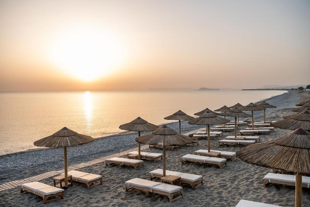 Pludmale ar saulrietu, kur redzami atpūtas krēsli un salmu saulessargi. Piekraste ir klāta ar smiltīm, un ūdens ir mierīgs un spīdīgs.