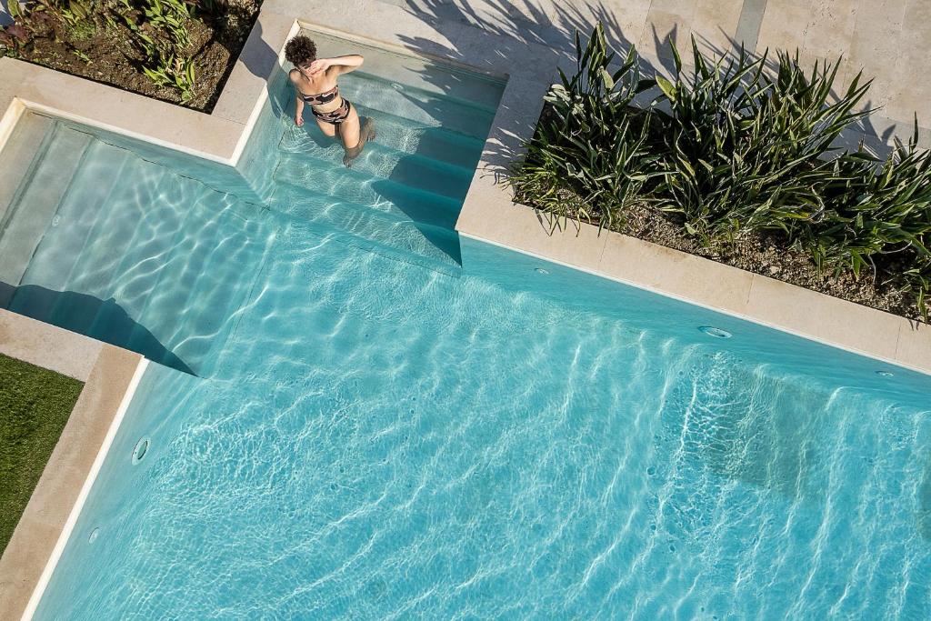 Peldbaseins ar skaidru ūdeni un augu apstādījumiem, kurā redzama persona, kas kāpj pa pakāpieniem. Ideāla vieta relaksācijai un atpūtai.