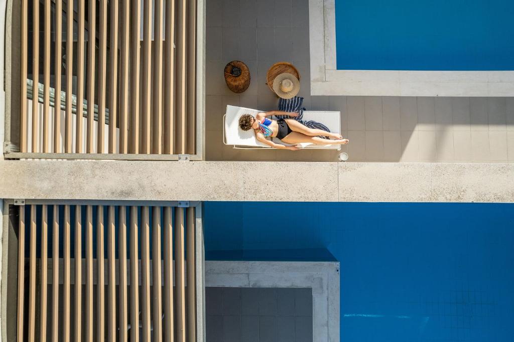 Saulainā diena pie baseina, kur sieviete atpūšas uz guļammaisa, baudot atpūtu un mieru. Apkārt redzami baseina zilie ūdeņi un mūsdienīga arhitektūra.