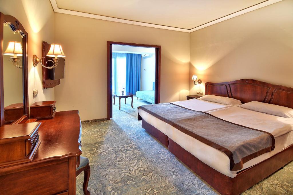 Mūsdienīga viesnīcas istaba ar diviem gultām, elegantu mēbelēm un dabīgu apgaismojumu. Telpā ir arī rakstāmgalds un liels logs, kas ved uz otro telpu.