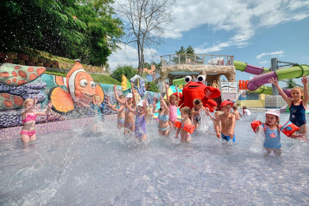 Bērni priecīgi spēlējas ūdens atrakciju parkā, izsistot ūdens strūklas un priecājoties ap krāsainām sienām ar jūras tēmu. Fons ietver ūdens slidkalniņus un saulainu debesi.