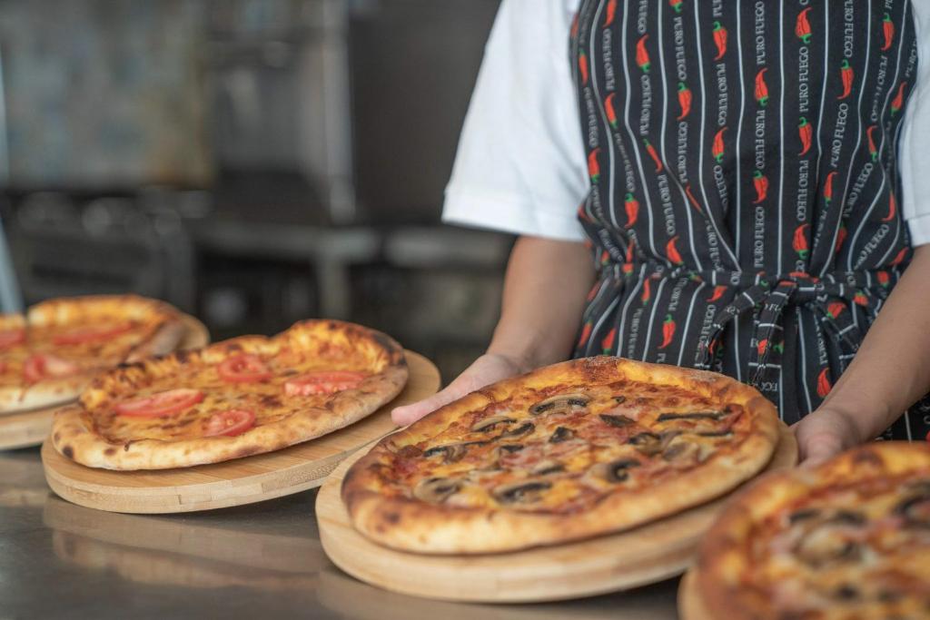 Picas pasniegšana restorānā, kur redzamas dažādas picas uz koka paletēm, un apkalpojošais personāls valkā priekšautu ar čili piparu motīvu.