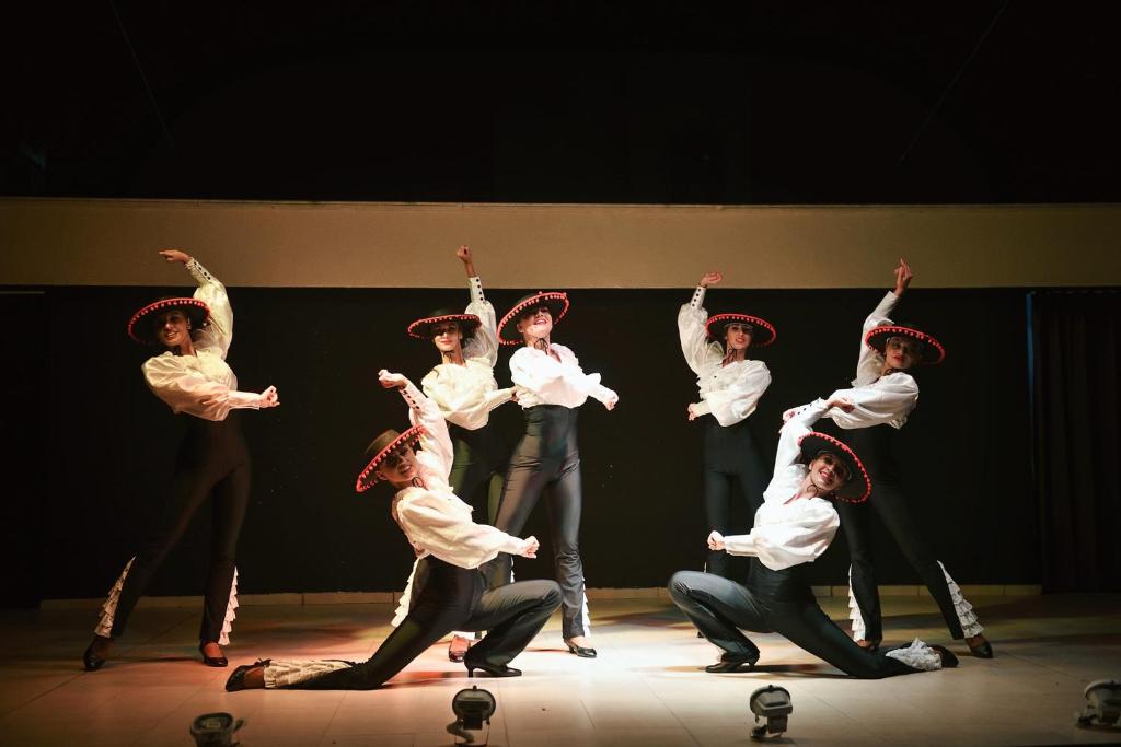 Deju grupa izpilda dinamisku uzstāšanos, tērpta baltos kreklos un melnās biksēs ar tradicionālajiem sombrero. Skatuve ir apgaismota ar mīkstu gaismu, radot radošu atmosfēru.