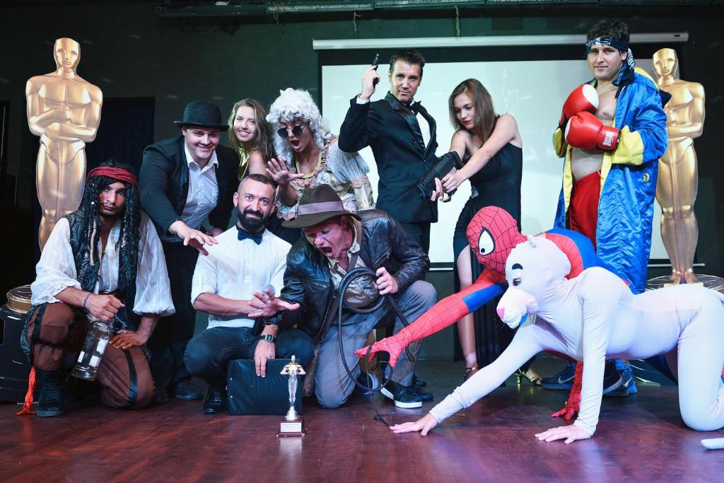 Grupas cilvēku, kas tērpušies dažādos tēlos, tostarp pirāts, supervaroņi un citi, pozē pie statuetes fona, kas atgādina Oskara balvu. Atmosfēra ir jautra un svinīga.