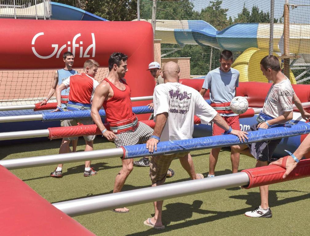 Cilvēki spēlē futbolu uz lielas, interaktīvas spēles platformas, kas imitē galda futbolu. Vides fons ir saulains, ar ūdens slidkalniņu.