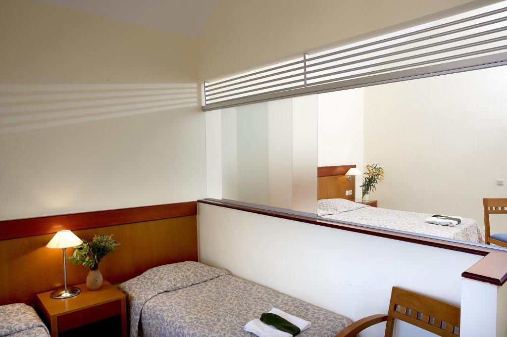 Gaiša guļamistaba ar divām gultām, naktsgaldiņu un lampu, kas rada mājīgu atmosfēru. Redzama arī spoguļa siena, kas paplašina telpas sajūtu.