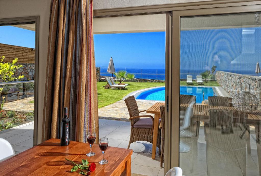 Elegantā viesnīcas istaba ar skatu uz baseinu un jūru, kur uz galda atrodas vīna pudele un glāzes. Dārza zona ar sauļošanās krēsliem un saulessargiem piepilda ainavu.