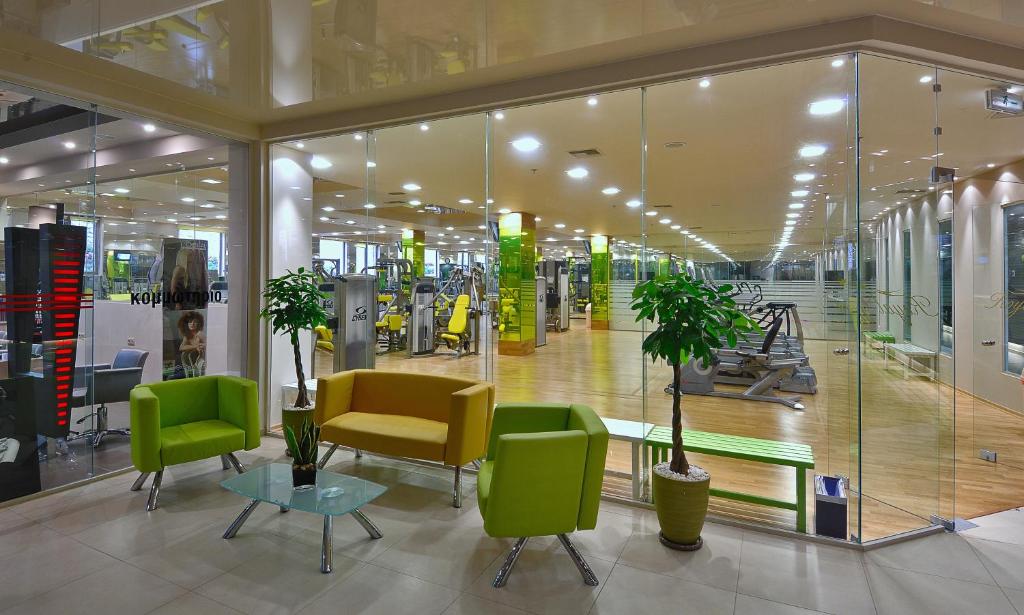 Moderns sporta zāles interjers ar stikla sienām, kurā redzami zaļas un dzeltenas krāsas krēsli, kafijas galdiņš un trenažieri. Telpa ir gaiša un plaša, radot aicinošu atmosfēru sportošanai.