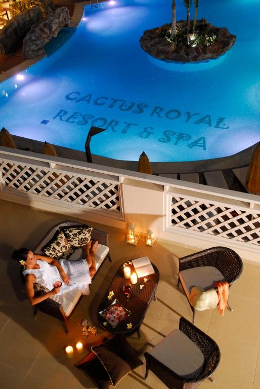 Relaksējoša aina pie baseina ar "Cactus Royal Resort & Spa" nosaukumu, kurā redzama sieviete, kas atpūšas uz krēsla. Apgaismojums rada mierīgu atmosfēru.