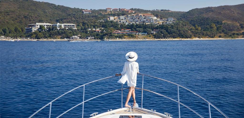 Sieviete ar baltu cepuri un apģērbu stāv uz jahtas priekša, vērojot skaistu piekrasti ar luksus mājām un zaļu dabu.