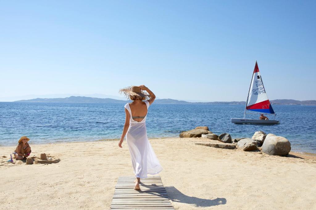 Sieviete baltā kleitā staigā pa pludmali, kamēr otra persona būvē smilšu pili. Jūras fons ar burājošu laivu un kalnainu ainavu.