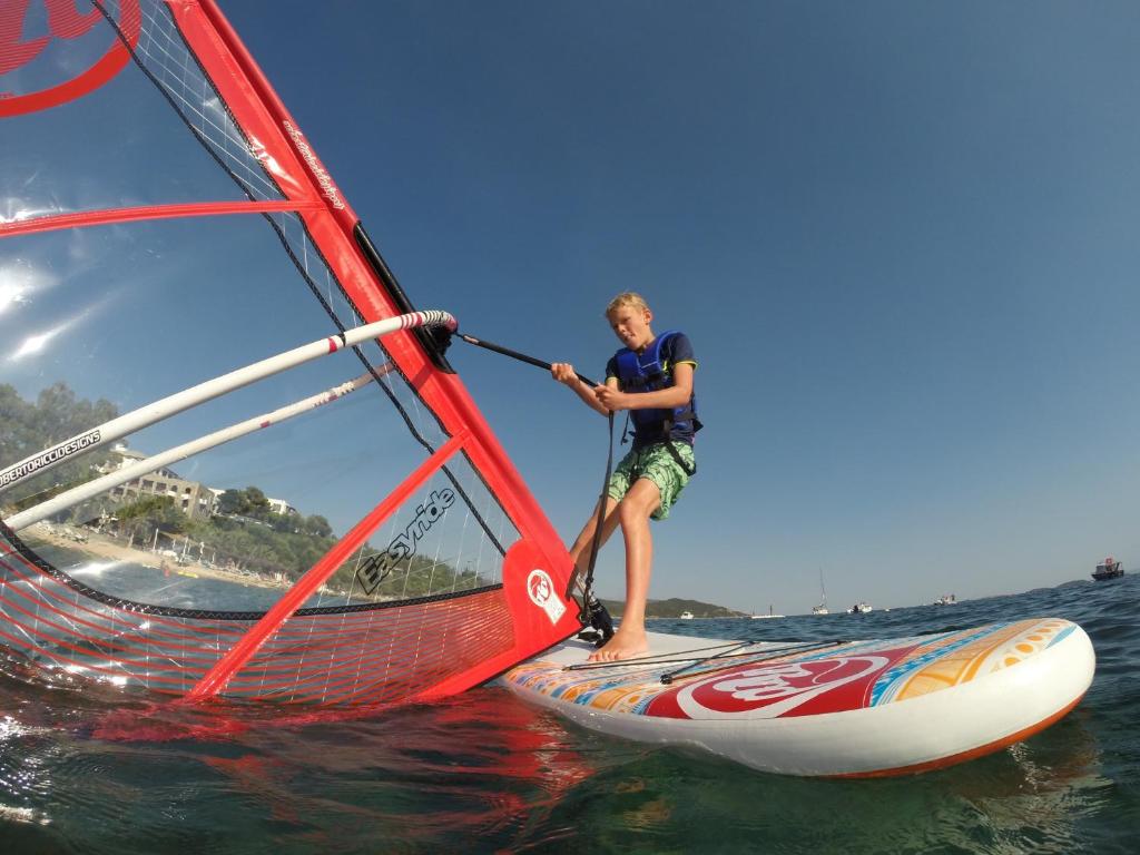 Jauns puisis, kas nodarbojas ar vindsērfingu, stāv uz dēļa ar sarkano buru, ūdens fona. Skats no apakšas uz burām un dēli, kas atspoguļo saulainu pludmales dienu.
