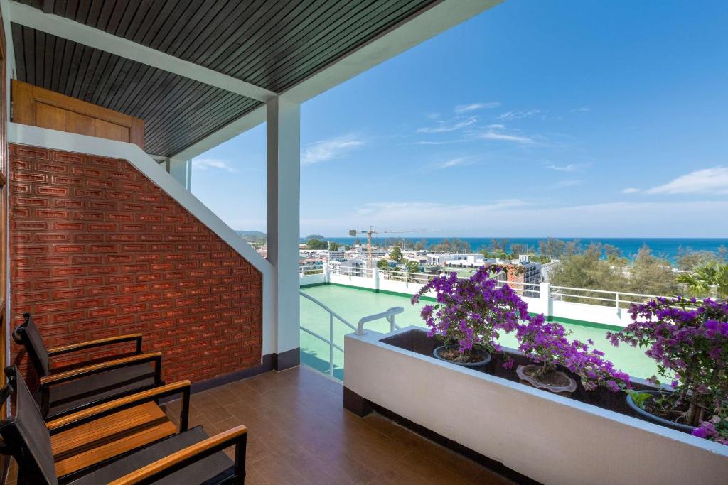 Balkons ar skatu uz jūru, kurā redzami divi koka krēsli un ziedi podos. Debesis ir skaidras un saulainas, radot relaksējošu atmosfēru.