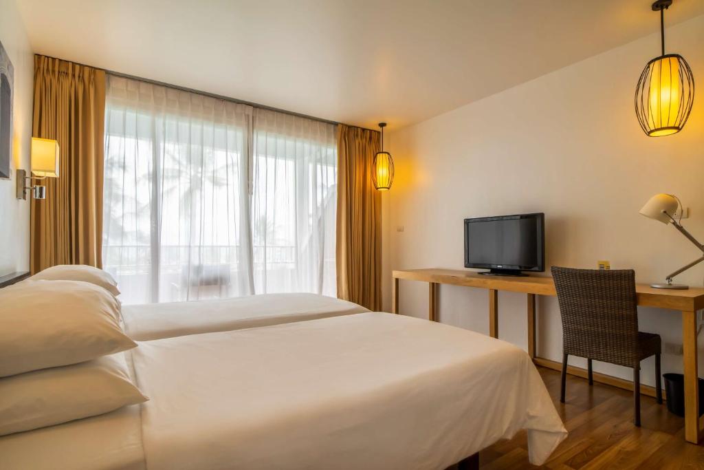 Mūsdienīga viesnīcas istaba ar divām gultām, televizoru un darba galdu, apgaismota ar stilīgām lampām un ar skatu uz terasi caur lieliem logiem.