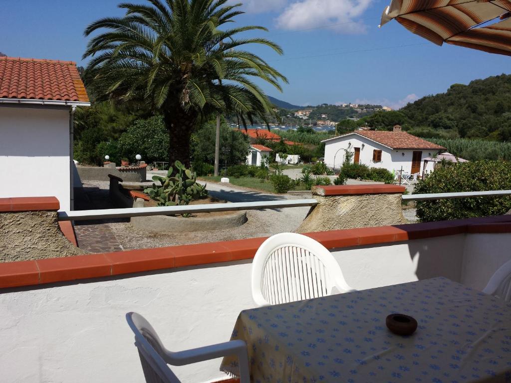 Atpūtas vieta ar skatu uz dārzu un palmu, kur pie galda ir zils galdauts. Fons veido gleznaini māju un kalnu ainavas ar skaidru debesu fonu.